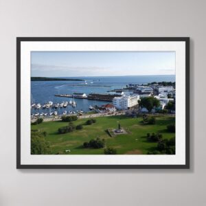 Aerial view of Mackinac Harbor and downtown on Mackinac Island, featuring a marina with sailboats, historic white buildings, green lawns, and the blue waters of Lake Huron under a sunny sky.