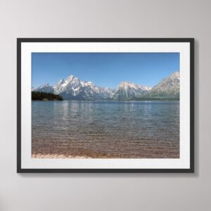 A scenic view of Jackson Lake at Grand Teton National Park with the majestic Teton mountain range reflected on the lake's crystal-clear waters and a rocky shoreline in the foreground.