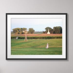 Photograph of a pioneer girl walking across the prairie at the Laura Ingalls Wilder Homestead in De Smet, South Dakota, with a historic church and windmill in the background.